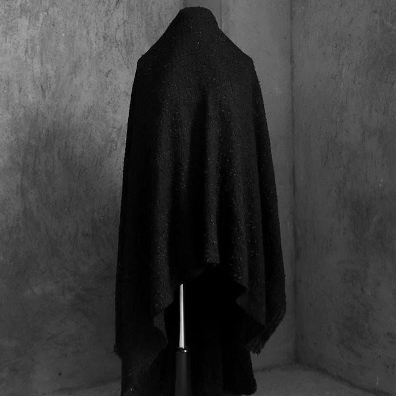 May include: A black, textured cloak draped over a mannequin. The cloak has a high collar and a flowing design, with a slightly rough, speckled surface. The background is a plain, gray wall.