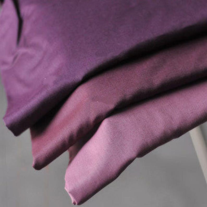 May include: Close-up of folded fabric in shades of purple, from deep violet to light lavender. The fabric appears soft and has a slight sheen, suggesting a luxurious material. The folds create shadows and highlights, adding depth to the image.