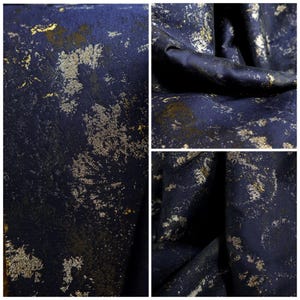 May include: Close-up of a dark blue fabric with a textured surface. The fabric has a pattern of gold and silver metallic flecks and splatters, creating a distressed, abstract design. The fabric is folded in some areas, showing depth and texture.
