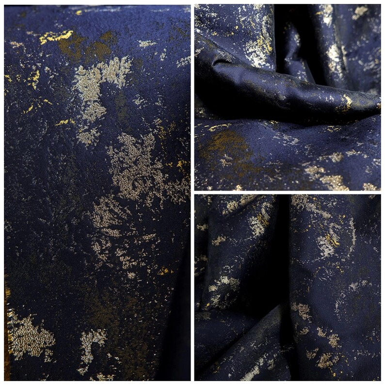 May include: Close-up of a dark blue fabric with a textured surface. The fabric has a pattern of gold and silver metallic flecks and splatters, creating a distressed, abstract design. The fabric is folded in some areas, showing depth and texture.