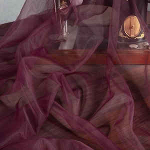 Peut inclure: Un tissu transparent bordeaux drapé sur une surface en bois, avec une machine à coudre vintage en arrière-plan. Le tissu a une texture délicate et fluide, et la couleur est une nuance riche de rouge-violet.