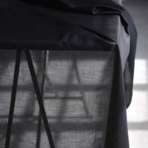 May include: A close-up shot of a black fabric draped over a table. The fabric has a smooth texture and is folded in a way that creates shadows and highlights. The table's metal frame is visible beneath the fabric, adding a geometric contrast to the soft folds.