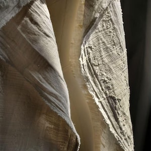 May include: Close-up of textured, light-colored fabric with visible weave and frayed edges. The fabric appears to be layered, with a lighter, smoother material visible beneath the textured outer layer. The lighting creates shadows and highlights the fabric's details.