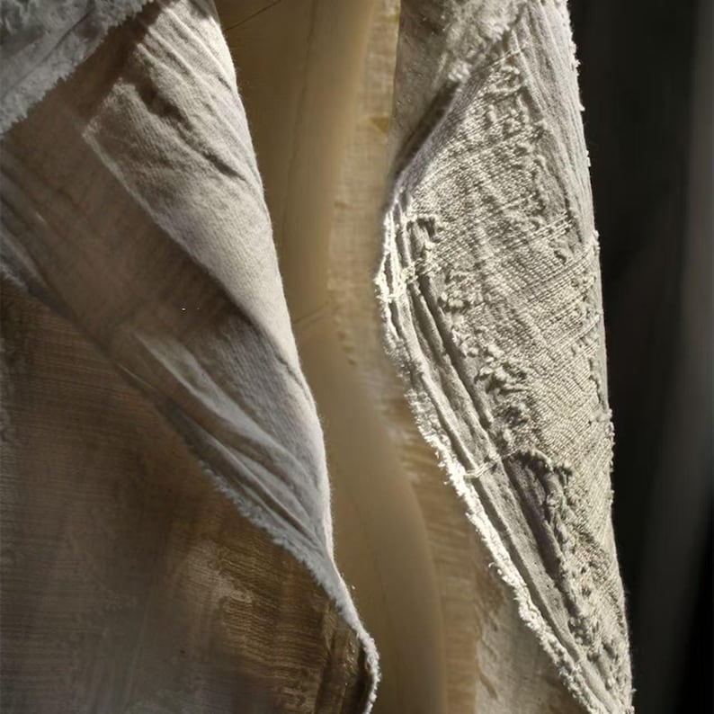 May include: Close-up of textured, light-colored fabric with visible weave and frayed edges. The fabric appears to be layered, with a lighter, smoother material visible beneath the textured outer layer. The lighting creates shadows and highlights the fabric's details.