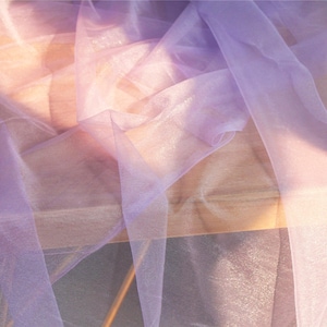Puede incluir: Una tela ligera y transparente en un suave tono lavanda con un brillo sutil. El material translúcido está drapeado en pliegues sueltos y fluidos, creando capas superpuestas que captan la luz y revelan una textura delicada y semitransparente.