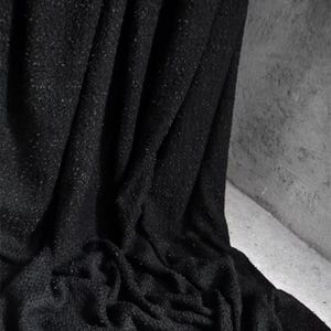 May include: A close-up shot of a black fabric with a textured surface. The fabric is draped, creating folds and shadows. The material appears to be soft and possibly woven, with small flecks of a lighter color throughout.