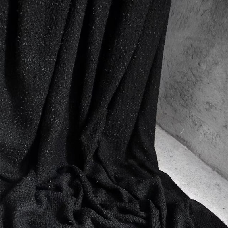 May include: A close-up shot of a black fabric with a textured surface. The fabric is draped, creating folds and shadows. The material appears to be soft and possibly woven, with small flecks of a lighter color throughout.