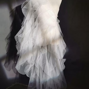 May include: A white, layered tulle fabric, possibly for a dress or skirt, is draped over a form. The fabric cascades down in multiple layers, creating a ruffled effect. The background is a dark gray, providing contrast to the bright white tulle.