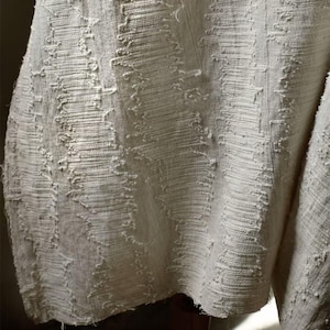 May include: Close-up of a textured, off-white fabric. The fabric has a woven appearance with a pattern of vertical and diagonal lines, creating a rough, tactile surface. The edges of the fabric appear unfinished, adding to its rustic aesthetic. The overall look is neutral and earthy.