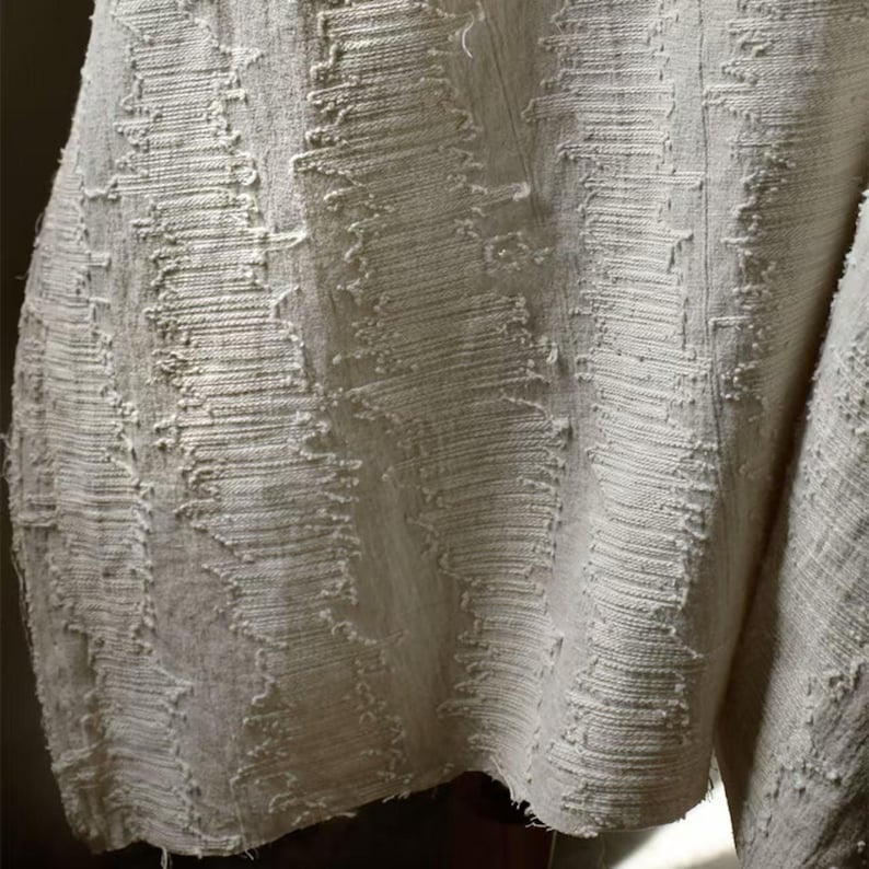 May include: Close-up of a textured, off-white fabric. The fabric has a woven appearance with a pattern of vertical and diagonal lines, creating a rough, tactile surface. The edges of the fabric appear unfinished, adding to its rustic aesthetic. The overall look is neutral and earthy.