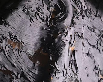 May include: Close-up of a textured, dark gray fabric with a layered, rippled design. The fabric has a three-dimensional quality, with raised sections and a flowing, organic pattern. The material appears to be soft and possibly silk or a similar drapeable textile.