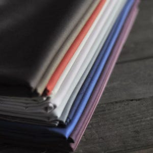 May include: A stack of folded fabric in various colors, including black, gray, red, white, blue, and purple. The fabrics are neatly arranged, showcasing a range of hues and textures. The image is taken on a dark wooden surface.