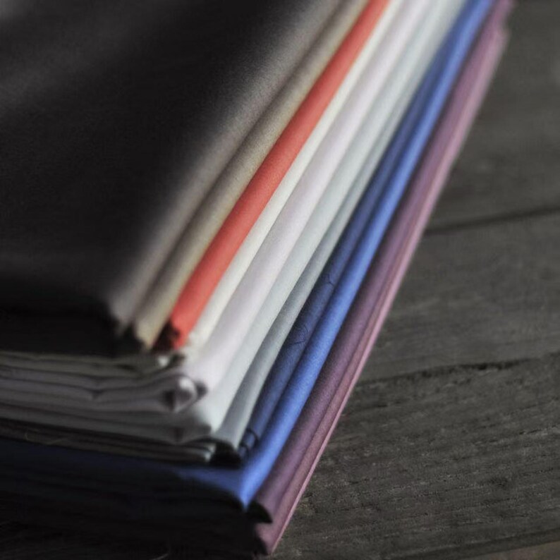 May include: A stack of folded fabric in various colors, including black, gray, red, white, blue, and purple. The fabrics are neatly arranged, showcasing a range of hues and textures. The image is taken on a dark wooden surface.