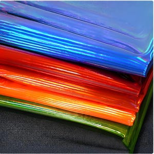 Könnte beinhalten: Ein Stapel schillernder Blätter in leuchtenden Farben: Blau, Rot, Orange und Grün. Die Blätter haben eine glänzende, reflektierende Oberfläche, die einen schimmernden Effekt erzeugt. Das Material scheint flexibel und geschichtet zu sein, mit einem dunklen, strukturierten Hintergrund.