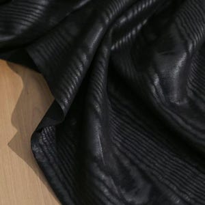 Puede incluir: Una pieza de tela negra con una sutil textura acanalada horizontal en relieve. El material est&aacute; drapeado para crear pliegues y sombras suaves, resaltando el brillo y el patr&oacute;n lineal constante en toda la superficie oscura.