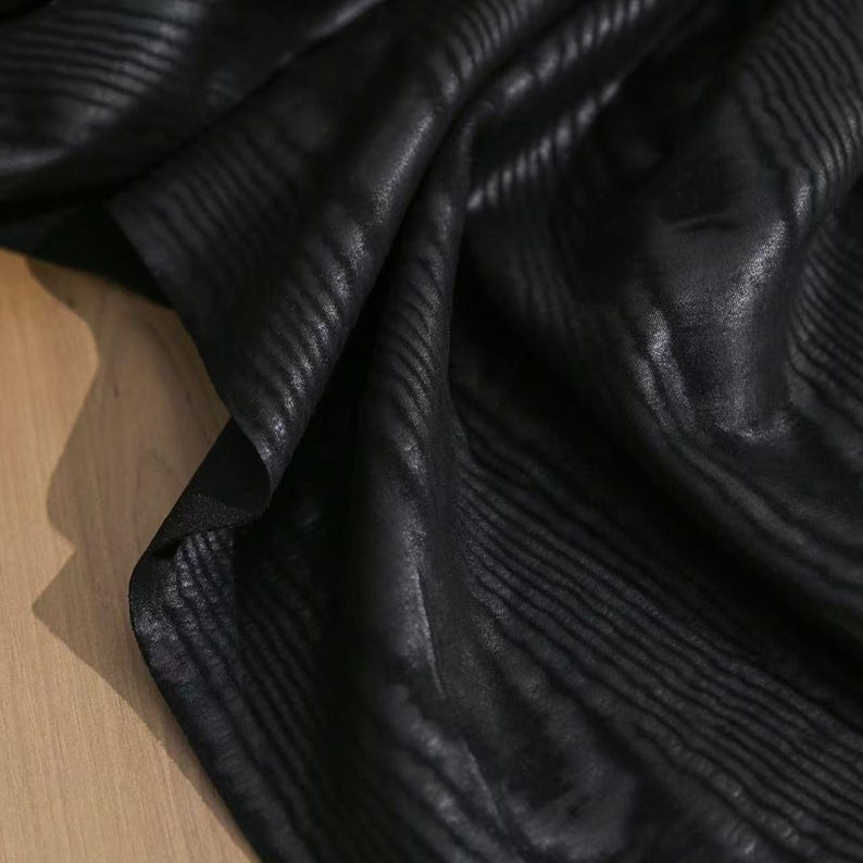 Puede incluir: Una pieza de tela negra con una sutil textura acanalada horizontal en relieve. El material est&aacute; drapeado para crear pliegues y sombras suaves, resaltando el brillo y el patr&oacute;n lineal constante en toda la superficie oscura.