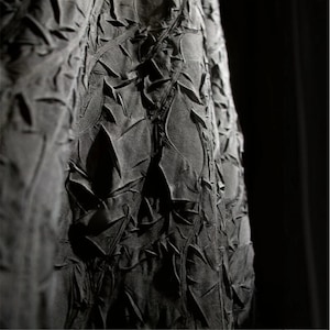 May include: Close-up of a dark gray fabric with a textured, layered design. The fabric appears to be folded and sculpted, creating a pattern of irregular shapes and shadows. The overall effect is a dramatic, artistic textile.