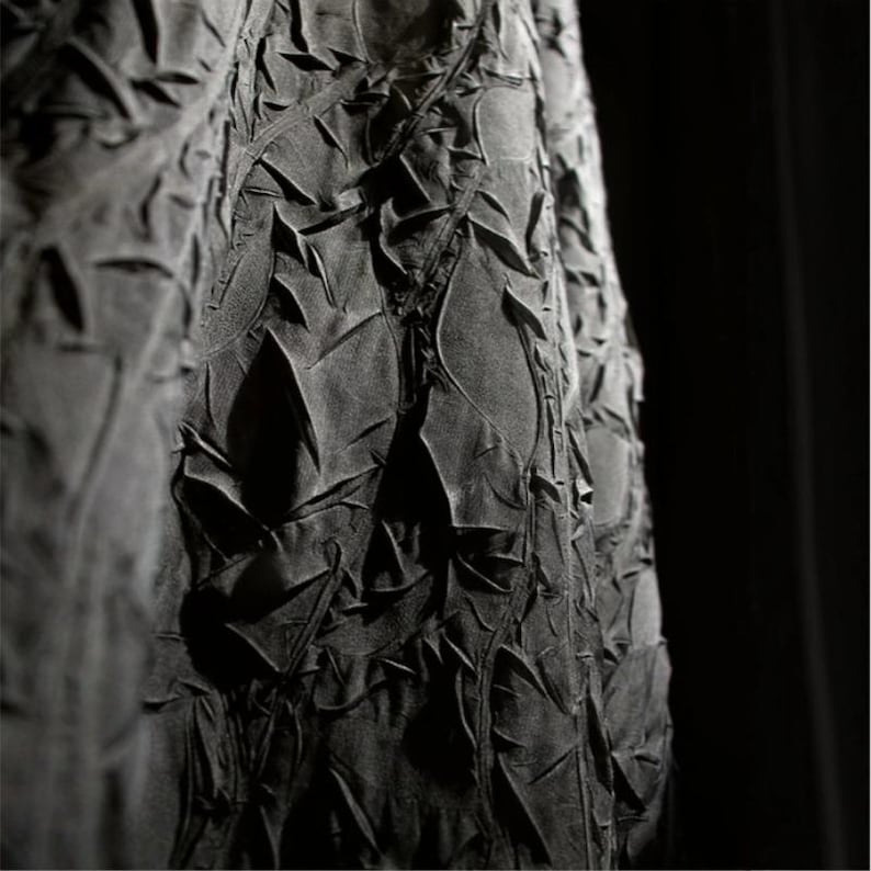 May include: Close-up of a dark gray fabric with a textured, layered design. The fabric appears to be folded and sculpted, creating a pattern of irregular shapes and shadows. The overall effect is a dramatic, artistic textile.