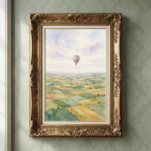 Könnte beinhalten: Ein gerahmtes Aquarellbild, das einen einzelnen Heißluftballon zeigt, der in einem blassblauen Himmel über einer weiten Landschaft aus grünen und gelben, patchworkartigen Feldern schwebt. Das Kunstwerk befindet sich in einem verzierten, goldfarbenen Rahmen.