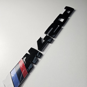 May include: Black M235i emblem with a red, dark blue, and light blue stripe design. The emblem is on a white background. The emblem is a car accessory.