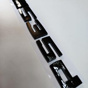 Puede incluir: Emblema negro brillante "335i" con una franja azul, roja y negra en la parte superior. Las letras gruesas y reflectantes destacan sobre un fondo blanco.