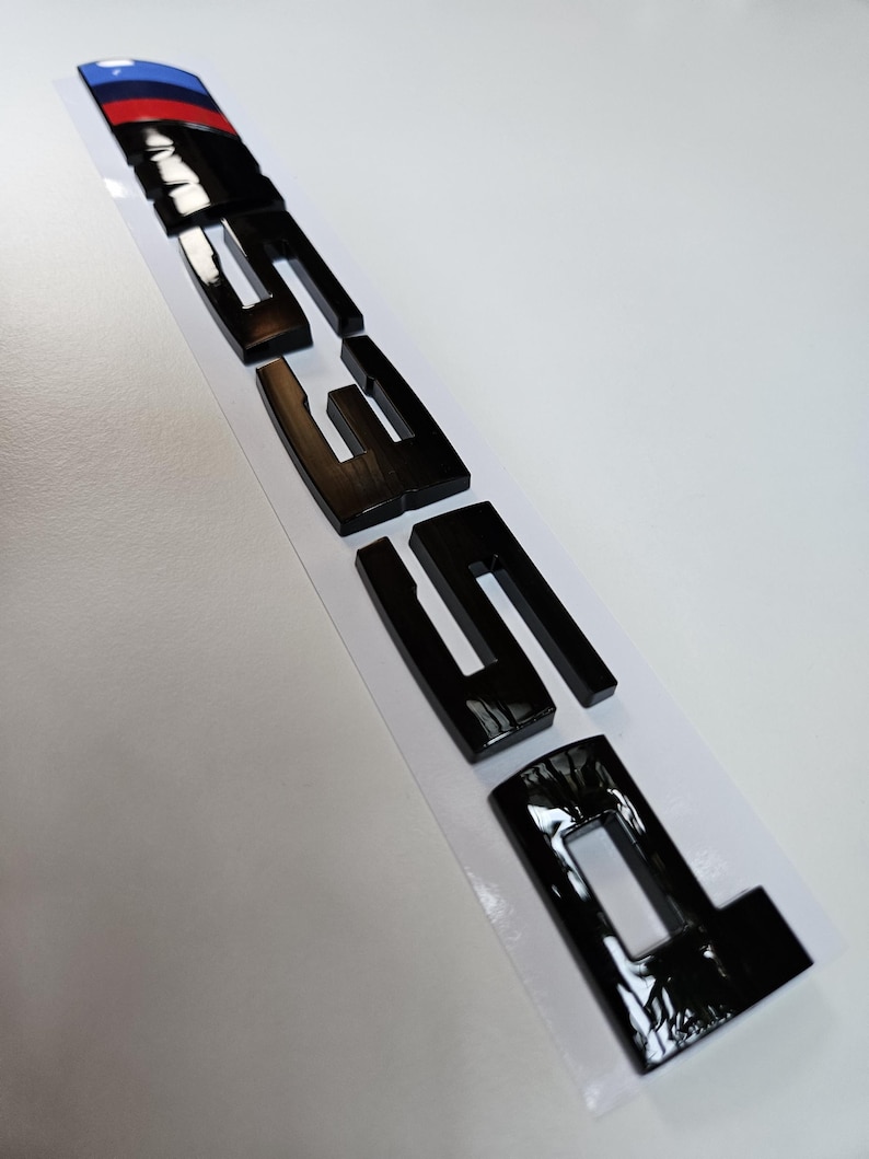 Puede incluir: Emblema negro brillante "335i" con una franja azul, roja y negra en la parte superior. Las letras gruesas y reflectantes destacan sobre un fondo blanco.