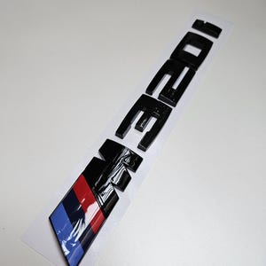 May include: Black car emblem with the text "E36 320i" in a glossy finish. The emblem features a colour stripe design with red, dark blue, and light blue stripes. The emblem is on a white backing.