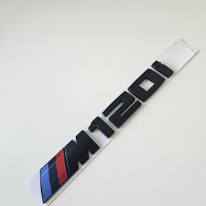 Op de afbeelding: Zwart M120i-embleem met blauwe, rode en zwarte strepen op een witte achtergrond. Het embleem is een auto-accessoire, waarschijnlijk voor een BMW-voertuig. De tekst M120i is vetgedrukt en zonder schreef.