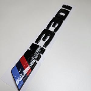 May include: Black "M30i" emblem with a color bar of blue, red, and black. The emblem is on a white backing. The letters are thick and glossy.