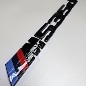 Puede incluir: Un emblema de coche negro brillante con las letras "M35i" y un dise&ntilde;o de bloques de color azul, rojo y negro. El emblema est&aacute; sobre un fondo blanco.