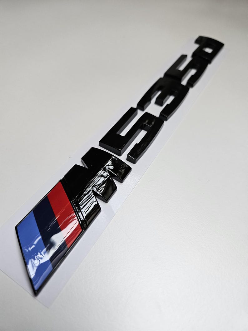 Puede incluir: Un emblema de coche negro brillante con las letras "M35i" y un dise&ntilde;o de bloques de color azul, rojo y negro. El emblema est&aacute; sobre un fondo blanco.