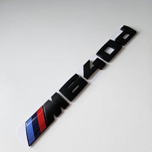 Pode incluir: Emblema M640i preto com um design de listras azuis, vermelhas e brancas. O emblema está em um fundo branco. As letras e os números estão em negrito, em uma fonte sem serifa.