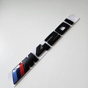 May include: Black M420i emblem with a blue, red, and white stripe design. The emblem is on a white backing. The letters and numbers are in a bold, sans-serif font.