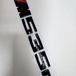 Puede incluir: Emblema de coche negro brillante con las letras "M550" y un dise&ntilde;o de rayas de color azul, rojo y negro. El emblema est&aacute; sobre un respaldo blanco, listo para su aplicaci&oacute;n. Accesorio automotriz.