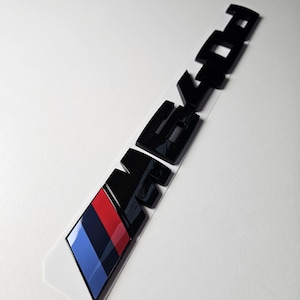 Pode incluir: Emblema M Performance preto com riscas vermelhas, azul escuro e azul claro. O emblema está sobre um suporte branco. As letras são arrojadas e brilhantes. O emblema está inclinado diagonalmente.