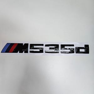 Puede incluir: Emblema negro M535d con el logotipo de la franja M de BMW en azul, rojo y azul oscuro. El emblema est&aacute; sobre un fondo blanco.