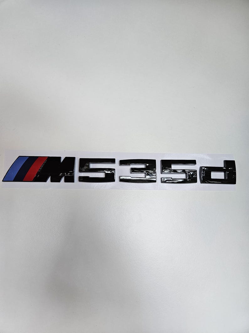 Puede incluir: Emblema negro M535d con el logotipo de la franja M de BMW en azul, rojo y azul oscuro. El emblema est&aacute; sobre un fondo blanco.