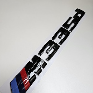 May include: Black M3 emblem with a glossy finish. The emblem features a colour bar with blue, red, and black stripes. The letters are bold and three-dimensional, designed to be applied to a vehicle.