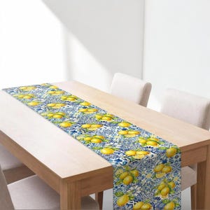 May include: A rectangular table runner with a vibrant lemon and blue tile pattern. The runner features yellow lemons and green leaves against a blue and white background. The runner is placed on a light-colored wooden table.