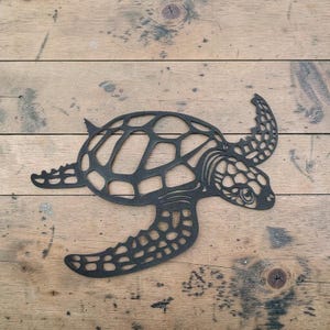 May include: A black metal sea turtle wall art piece. The turtle has a detailed openwork design, with a patterned shell and flippers. The artwork is placed on a wooden surface.