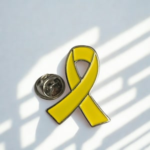 Puede incluir: Broche de lazo amarillo con respaldo plateado. El broche tiene forma de lazo anudado, un símbolo de apoyo y concienciación. El broche está sobre un fondo blanco con sombras.