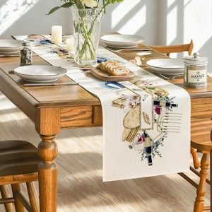 May include: A cream-coloured table runner with a watercolour design featuring wine bottles, cheese, and books. The runner is placed on a wooden table set for a meal, with plates, silverware, and a vase of flowers.