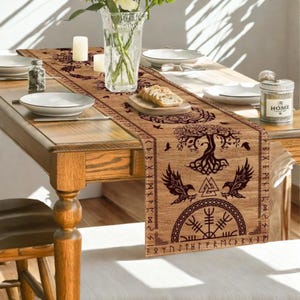 Può includere: Runner da tavolo marrone con un design a tema vichingo con un albero, corvi e simboli runici. Il runner è posto su un tavolo di legno apparecchiato per un pasto, con piatti, candele e un vaso di fiori.
