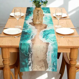 May include: A rectangular table runner with a watercolour design of a beach scene. The runner features turquoise waves, a sandy shore, and a light brown background. The runner is placed on a wooden table set with plates, glasses, and cutlery.