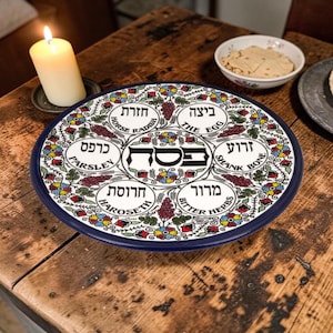 Peut inclure: Une assiette de Seder en céramique avec un bord bleu, présentant du texte hébreu et des traductions anglaises. L'assiette est ornée de motifs floraux et de raisins rouges, jaunes et verts. Une bougie allumée et un petit bol de nourriture sont visibles.