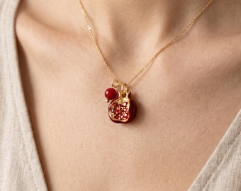 May include: A gold-tone chain necklace featuring a pomegranate-shaped pendant with deep red enamel and small red crystals, accompanied by a single round red bead charm hanging from the same gold-tone jump ring.