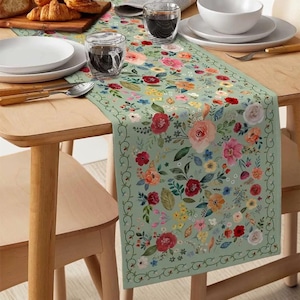 May include: A floral table runner with a light green background and a colorful pattern of flowers in shades of red, pink, yellow, and blue. The runner is rectangular and is placed on a wooden table, with plates, glasses, and cutlery.
