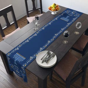 May include: A navy blue table runner with a decorative border and menorah design. The runner is placed on a dark wooden table set for a meal, with plates, silverware, and wine glasses.