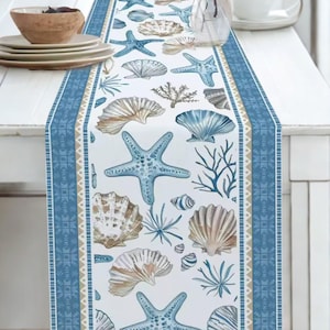 Könnte beinhalten: Ein weißer Tischläufer mit einem sich wiederholenden Muster aus blauen Seesternen, verschiedenen Muscheln und Korallenzweigen. Der Läufer ist mit einem blauen geometrischen Druck und dünnen beigen Streifen eingefasst. Das Design ist auf Textil.