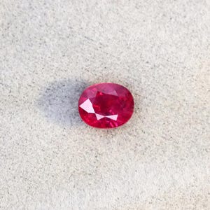 Könnte beinhalten: Ein facettierter, oval geformter Rubin-Edelstein. Der Edelstein hat eine tiefe, leuchtende rote Farbe mit mehreren Facetten, die das Licht reflektieren. Der Rubin liegt auf einer hellgrauen, strukturierten Oberfläche.